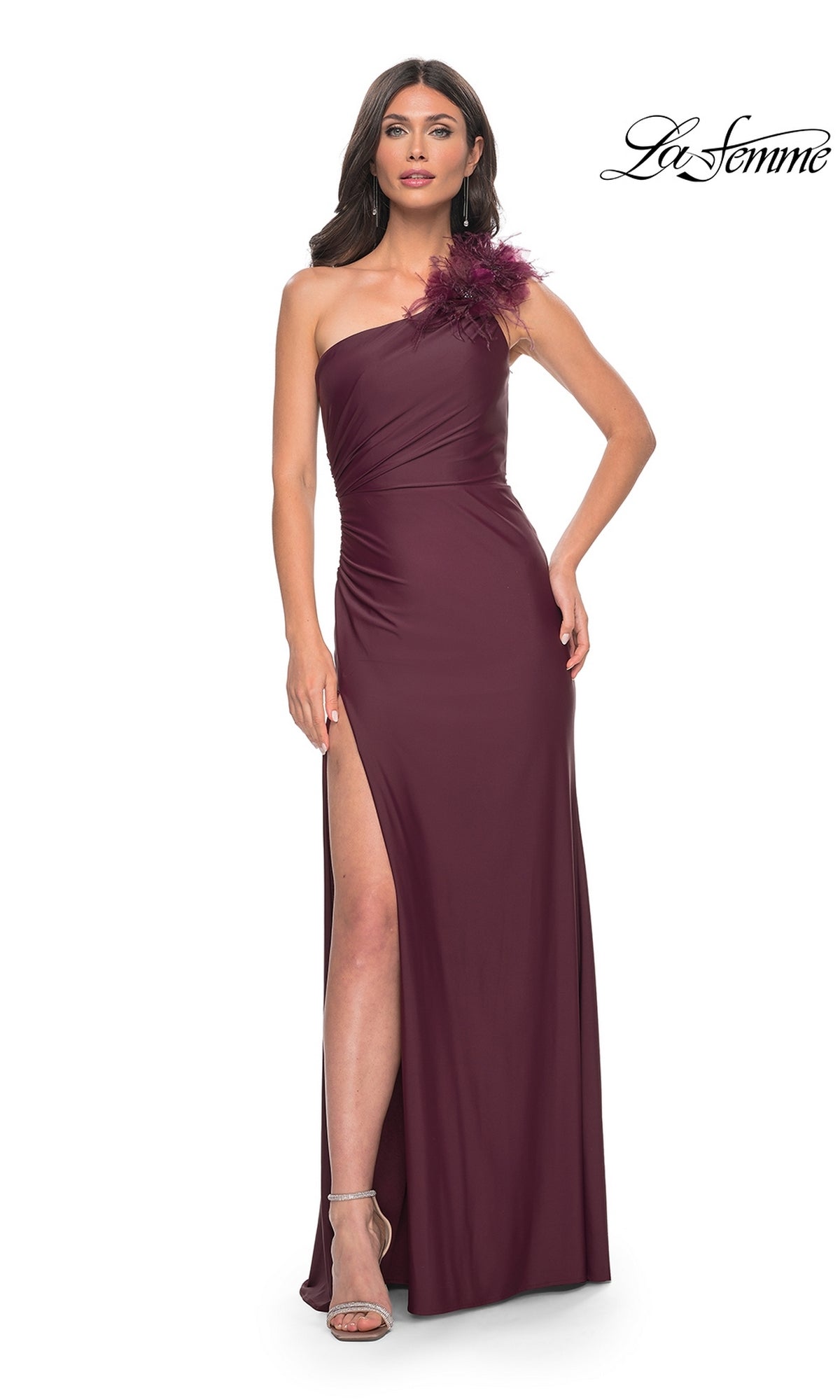 A woman models the La Femme Feathered One-Shoulder Prom Dress 32076 in dark wine, featuring feather detail and a thigh-high slit, paired with silver heels against a white background.
